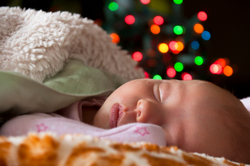 A baby sleeps with christmas lights behind them.